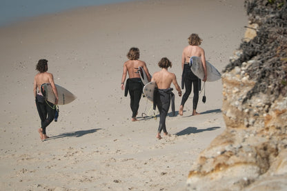 A group of four surfers walking on a sandy beach carrying their surfboards, with the ocean and rocks in the background.